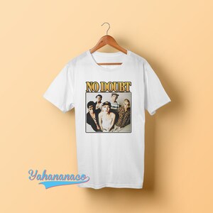 May include: White t-shirt with a vintage-style graphic of the band No Doubt. The graphic features a photo of the band members, including Gwen Stefani, Tony Kanal, Adrian Young, Tom Dumont, and  the band's name in gold letters.