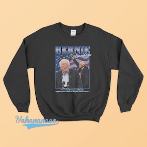 Pode incluir: Camisola de moletom preta com um gráfico de estilo vintage de Bernie Sanders. O gráfico apresenta uma foto de Sanders com o texto "Bernie Sanders" e uma citação: "You can't become a billionaire stepping over children sleeping on the street."