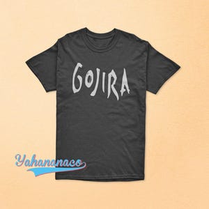 Op de afbeelding: Een zwart t-shirt met korte mouwen en het woord GOJIRA in een lichtgrijs, gestileerd en grillig lettertype op de borst gedrukt. Het shirt heeft een ronde hals en een standaard pasvorm. De tekst Yahanaco is linksonder in beeld zichtbaar.