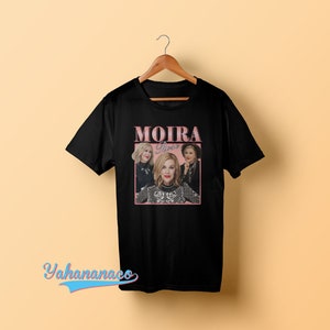 以下が含まれることがあります： 黒いTシャツに、ピンク色のグラフィックで3人の女性が描かれており、その下に「MOIRA Rose」という文字が書かれています。