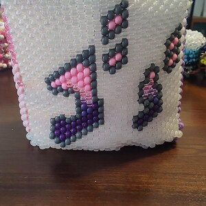 May include: A white beaded cube-shaped container with a musical note design. The notes are made of gray, pink, and purple beads. The container is sitting on a wooden surface.