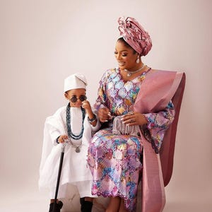 May include: A woman and child in traditional Nigerian clothing are seated. The woman wears a patterned gown and head wrap. The child is in a white outfit with sunglasses and a cane. The image showcases cultural elegance, with vibrant colours and intricate patterns.