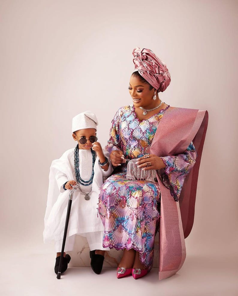 May include: A woman and child in traditional Nigerian clothing are seated. The woman wears a patterned gown and head wrap. The child is in a white outfit with sunglasses and a cane. The image showcases cultural elegance, with vibrant colours and intricate patterns.