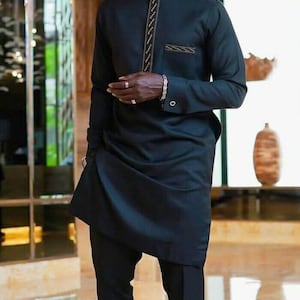 Könnte beinhalten: Schwarzes Tunika- und Hosen-Outfit. Die langärmlige Tunika hat einen Stehkragen und eine dekorative goldfarbene Verzierung auf der Vorderseite. Das Outfit beinhaltet eine passende schwarze Hose. Der Mann lächelt.