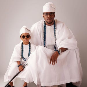 May include: Two people in matching white traditional attire, hats, and beaded necklaces. One wears glasses, the other sunglasses. The person on the left holds a cane and wears black shoes. The other wears black dress shoes.