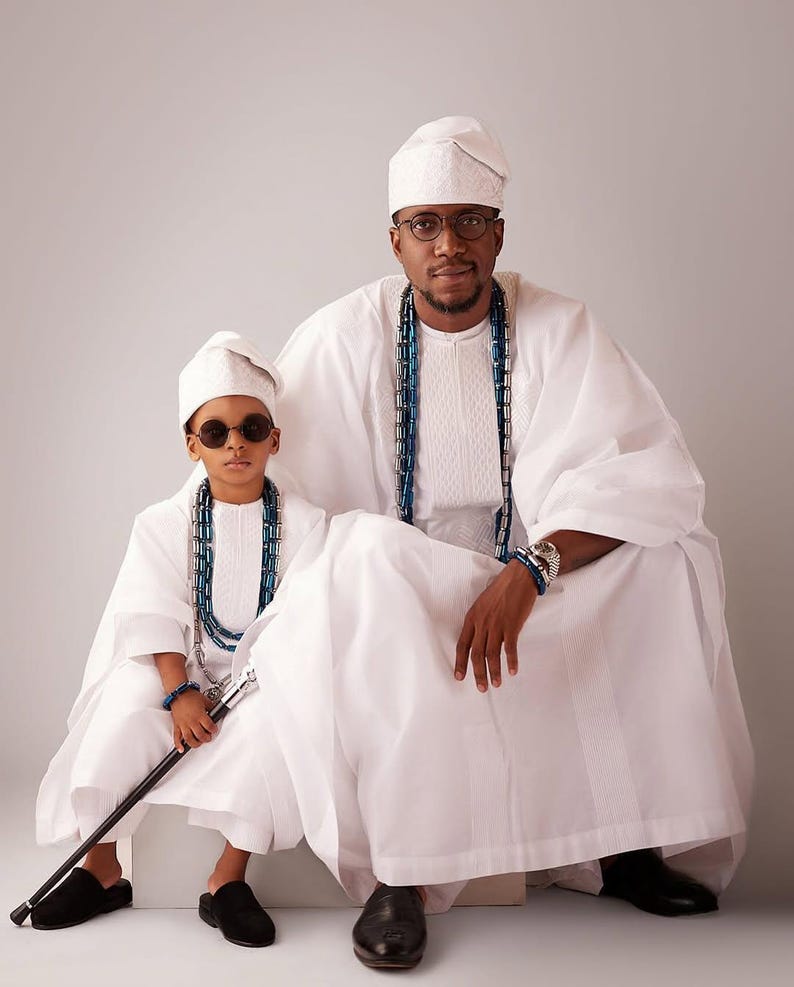 May include: Two people in matching white traditional attire, hats, and beaded necklaces. One wears glasses, the other sunglasses. The person on the left holds a cane and wears black shoes. The other wears black dress shoes.