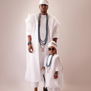 May include: Two people in traditional white clothing, including robes, hats, and beaded necklaces. The adult wears glasses and the child wears sunglasses. Both have bracelets and black shoes. The child holds a cane.