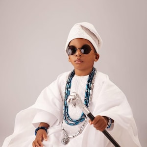 May include: A person in traditional white clothing, wearing round sunglasses and a white cap. They are adorned with a blue and silver beaded necklace and bracelet, and hold a silver-topped cane. The outfit is a cultural garment.