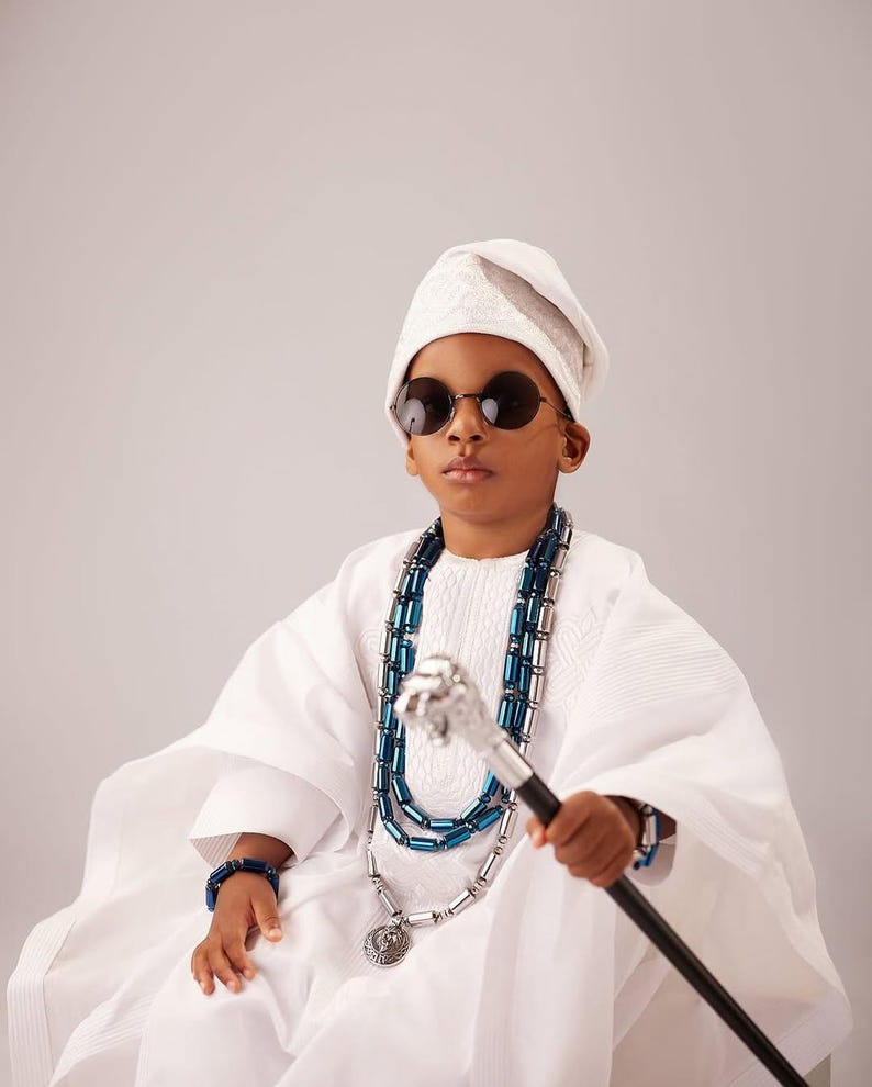 May include: A person in traditional white clothing, wearing round sunglasses and a white cap. They are adorned with a blue and silver beaded necklace and bracelet, and hold a silver-topped cane. The outfit is a cultural garment.