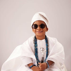 May include: A child in traditional white clothing, wearing round sunglasses and a white cap. The child is adorned with a blue and silver beaded necklace and bracelets, and holds a decorative cane with a silver top.