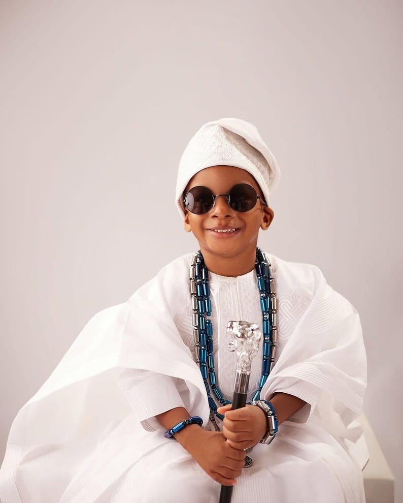 May include: A child in traditional white clothing, wearing round sunglasses and a white cap. The child is adorned with a blue and silver beaded necklace and bracelets, and holds a decorative cane with a silver top.