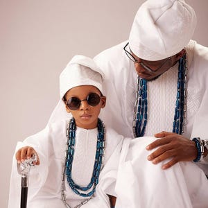 May include: Two people, possibly a father and child, are dressed in traditional white clothing. The child wears round sunglasses and holds a cane. Both wear beaded necklaces with blue and silver accents. They are wearing white hats.