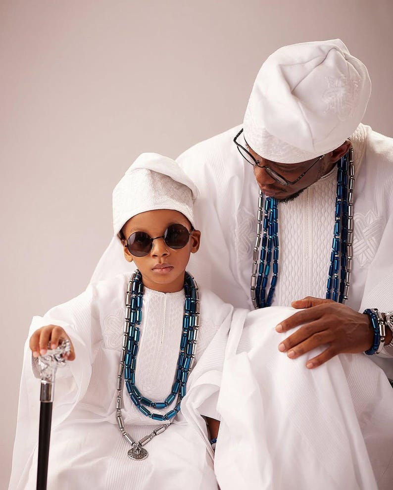 May include: Two people, possibly a father and child, are dressed in traditional white clothing. The child wears round sunglasses and holds a cane. Both wear beaded necklaces with blue and silver accents. They are wearing white hats.