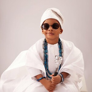 May include: A person in traditional white clothing, wearing round sunglasses and a white cap. They are adorned with a blue and silver beaded necklace and bracelets, and holding a decorative cane with a silver top. The outfit is a cultural representation.