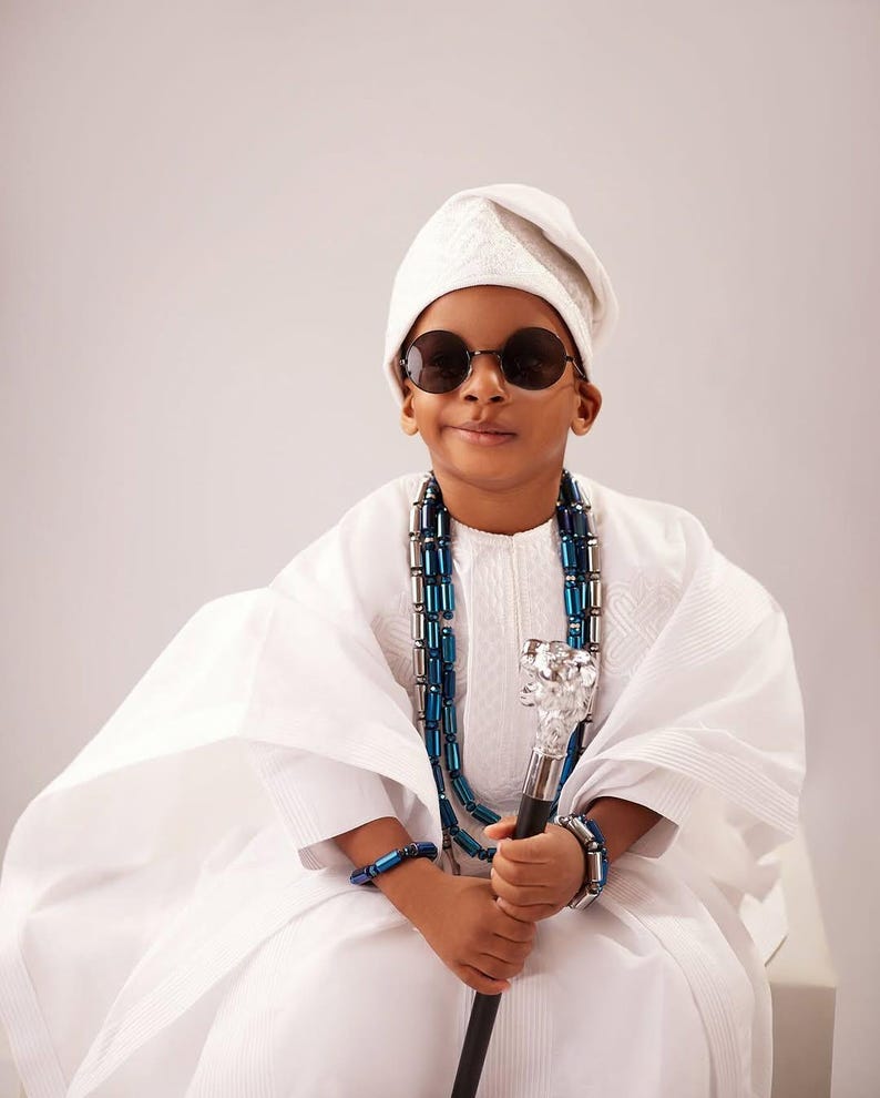 May include: A person in traditional white clothing, wearing round sunglasses and a white cap. They are adorned with a blue and silver beaded necklace and bracelets, and holding a decorative cane with a silver top. The outfit is a cultural representation.