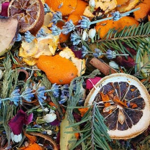 May include: A close-up of a bowl filled with dried citrus peels, lavender flowers, cinnamon sticks, and other natural ingredients. The mixture is a blend of orange, brown, green, and purple hues.