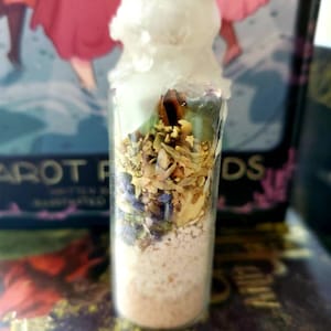 May include: A clear glass bottle with a white wax top filled with dried herbs, flowers, and a white powder. The bottle is sitting on a surface with a book in the background.