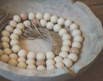 May include: A wooden bowl filled with a long strand of natural wood beads. The beads are strung together with twine and have a natural, light brown color. The bowl is made of wood and has a rustic, natural look.
