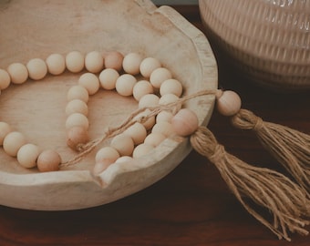 May include: A wooden bead garland with two tassels, the garland is in a wooden bowl, the tassels are made of natural fiber.