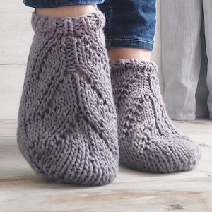Puede incluir: Un par de calcetines grises de punto con un patrón calado detallado. Los calcetines se usan con vaqueros azules. Los calcetines están hechos de un hilo suave y texturizado y tienen un puño acanalado.