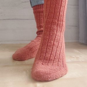 May include: Coral-coloured, ribbed knit socks. The knee-high socks have a textured pattern and are worn with blue denim jeans, cuffed at the ankles. The background is a light wood-panelled wall. The socks are made of a soft material.