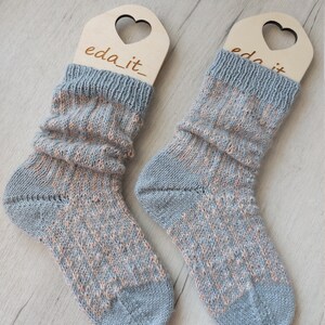 May include: A pair of knitted socks in blue and peach hues, presented on wooden heart-shaped hangers. The socks feature a textured design and a ribbed cuff. The text "eda_it" is visible on the hangers.