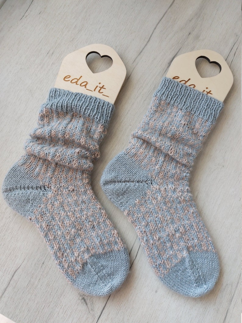 May include: A pair of knitted socks in blue and peach hues, presented on wooden heart-shaped hangers. The socks feature a textured design and a ribbed cuff. The text "eda_it" is visible on the hangers.