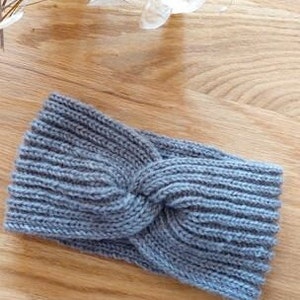 May include: A gray knitted headband with a twisted knot design. The headband is made of a ribbed knit fabric and is lying on a wooden surface. The headband is a solid color and appears to be handmade.