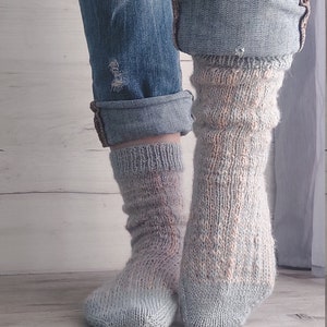 May include: A pair of handmade, light blue and peach-coloured knitted socks. The socks are worn with rolled-up denim jeans. The socks have a textured, ribbed design and are made of soft yarn.