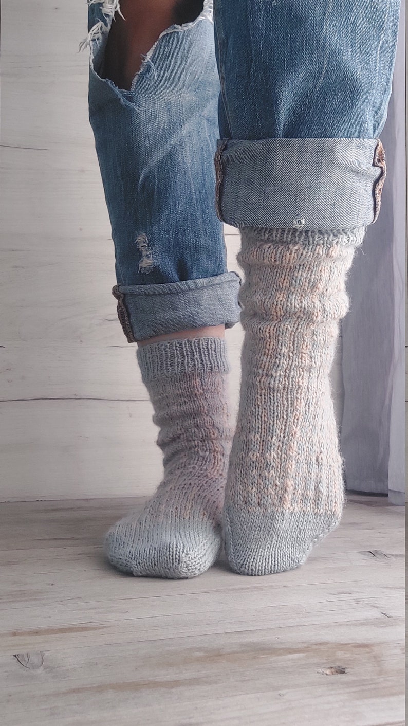 May include: A pair of handmade, light blue and peach-coloured knitted socks. The socks are worn with rolled-up denim jeans. The socks have a textured, ribbed design and are made of soft yarn.
