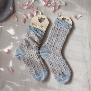 May include: Two knitted socks in pale blue and peach, featuring a ribbed design. They are displayed on wooden hangers with heart-shaped cutouts. The text "eda_it_" is visible on the hangers. Pink flower petals are scattered around the socks.