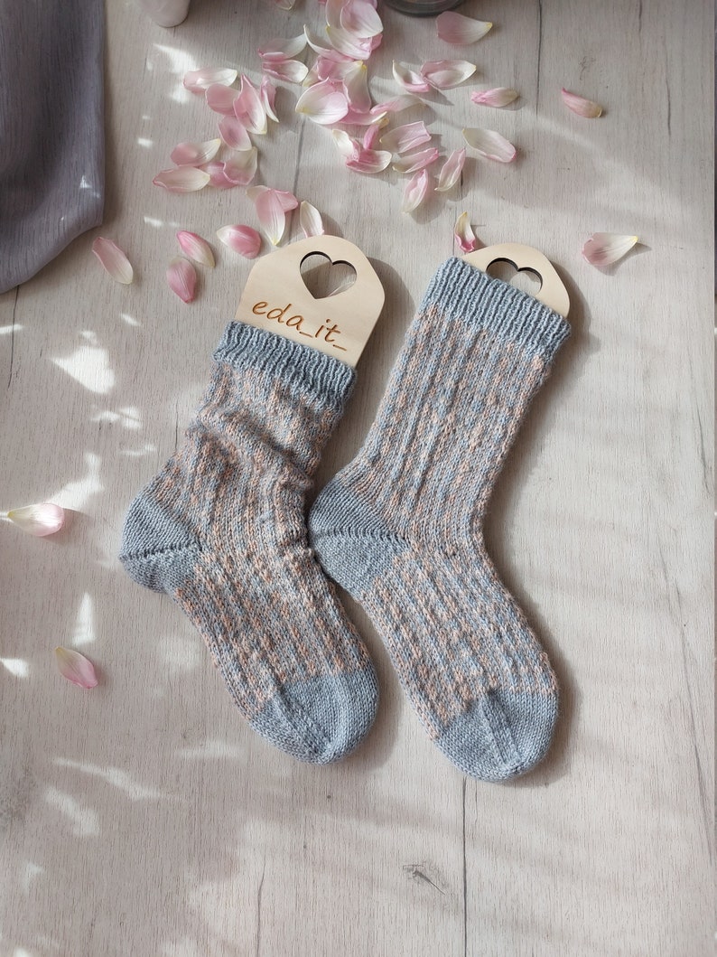 May include: Two knitted socks in pale blue and peach, featuring a ribbed design. They are displayed on wooden hangers with heart-shaped cutouts. The text "eda_it_" is visible on the hangers. Pink flower petals are scattered around the socks.