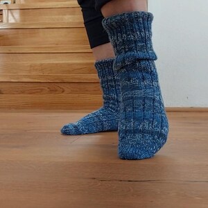 Puede incluir: Un par de calcetines azules de punto con un patr&oacute;n texturizado. Los calcetines se llevan con pantalones oscuros y est&aacute;n sobre un suelo de madera. Los calcetines son una mezcla de hilos azules y blancos.