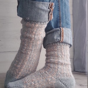 May include: Knitted socks in blue and peach tones, paired with rolled-up denim jeans. The socks feature a textured design and are visible from the ankles down, set against a wooden backdrop.