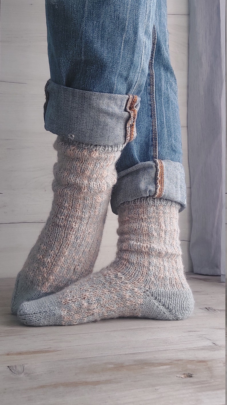 May include: Knitted socks in blue and peach tones, paired with rolled-up denim jeans. The socks feature a textured design and are visible from the ankles down, set against a wooden backdrop.