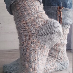 May include: Hand-knitted socks in light blue, peach, and cream. The socks have a textured pattern and are visible from the ankles down, with denim jeans folded above. The socks are made of soft yarn.