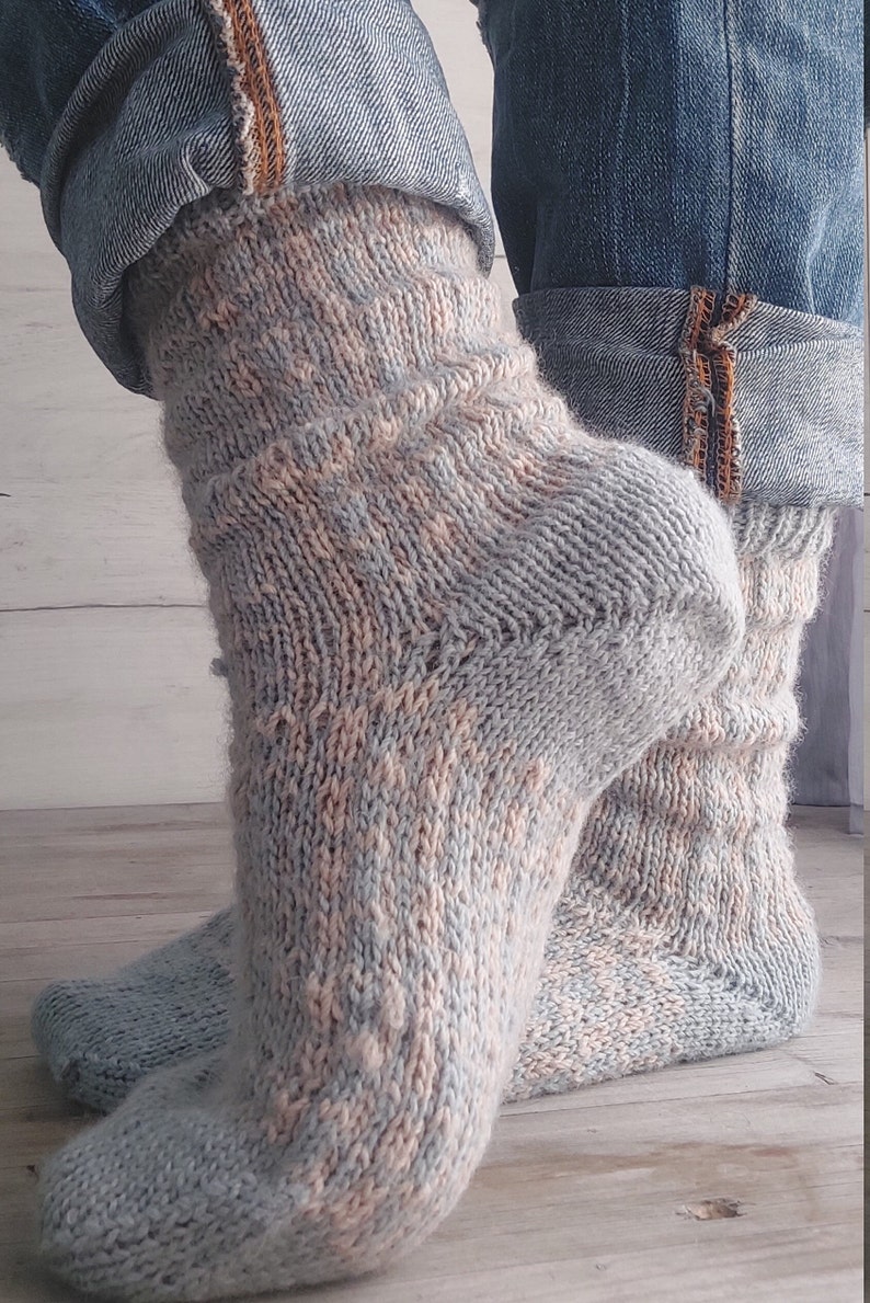May include: Hand-knitted socks in light blue, peach, and cream. The socks have a textured pattern and are visible from the ankles down, with denim jeans folded above. The socks are made of soft yarn.