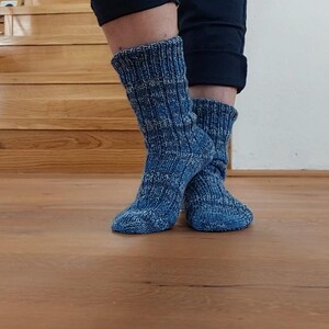 Puede incluir: Un par de calcetines azules de punto con un dise&ntilde;o acanalado. Los calcetines se llevan puestos, con las piernas y los pies visibles. Los calcetines son de color azul medio con un aspecto texturizado. El fondo incluye escaleras y suelo de madera.