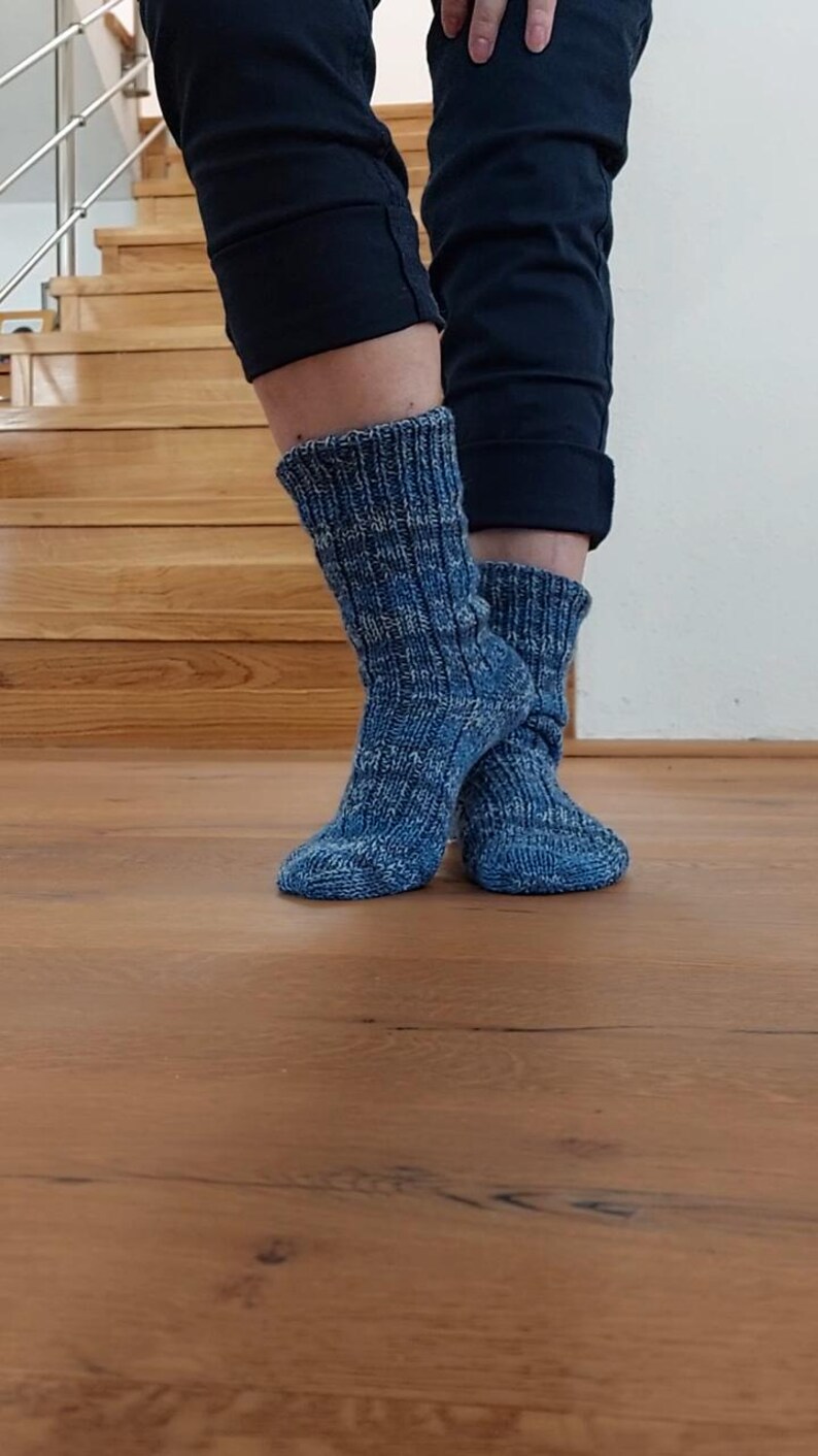 Puede incluir: Un par de calcetines azules de punto con un dise&ntilde;o acanalado. Los calcetines se llevan puestos, con las piernas y los pies visibles. Los calcetines son de color azul medio con un aspecto texturizado. El fondo incluye escaleras y suelo de madera.