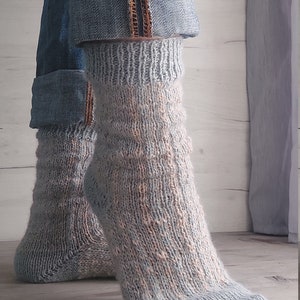 May include: A pair of light blue and peach-coloured knitted socks. The socks are worn with rolled-up denim jeans. The socks have a textured pattern and are made of a soft, warm material. The jeans have a distressed look.
