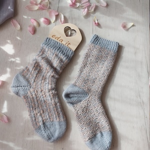 May include: A pair of hand-knitted socks in shades of blue, beige, and brown, with a ribbed cuff and toe. The socks are displayed on a light wooden surface, with a wooden tag that says "eda it." Pink flower petals are scattered around the socks.