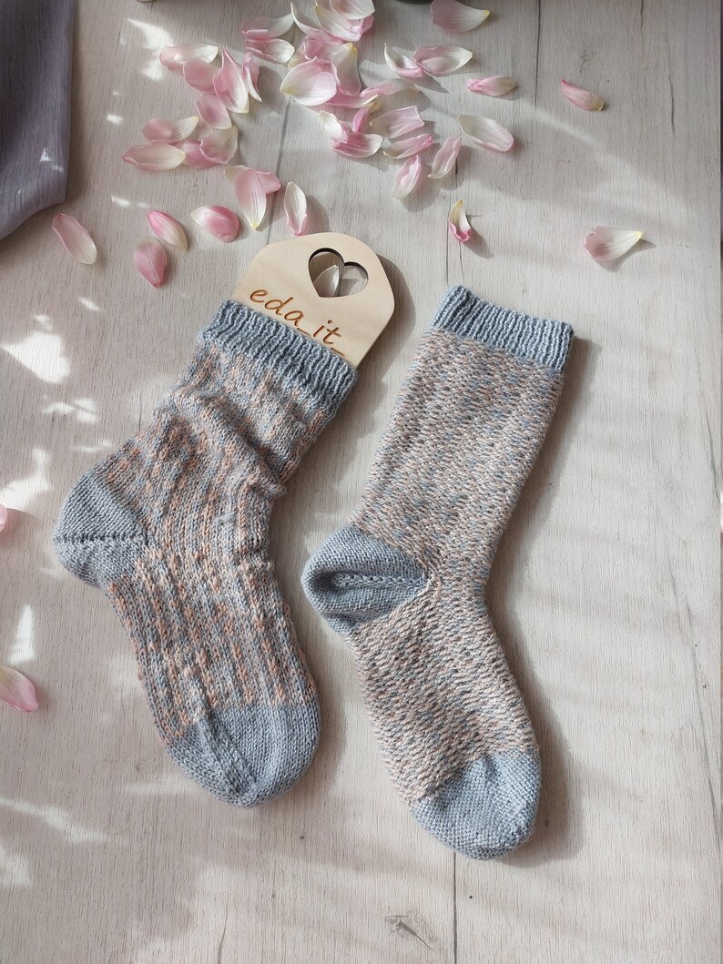 May include: A pair of hand-knitted socks in shades of blue, beige, and brown, with a ribbed cuff and toe. The socks are displayed on a light wooden surface, with a wooden tag that says "eda it." Pink flower petals are scattered around the socks.