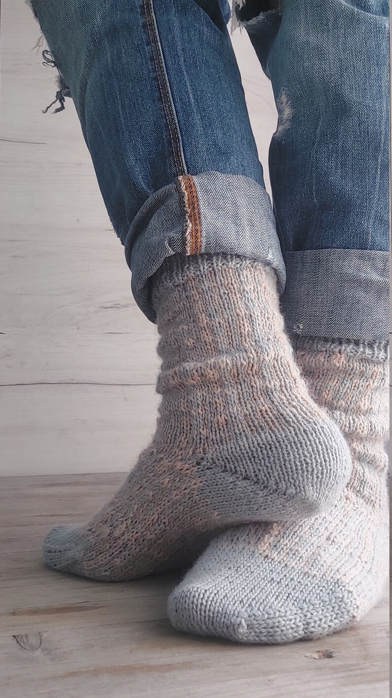 May include: A pair of light blue and peach-coloured knitted socks. The socks are worn with blue denim jeans, cuffed at the ankles. The socks have a textured pattern and are made of a soft, warm material.