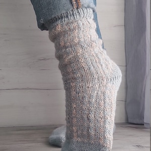 May include: A close-up of a pair of knitted socks in shades of light blue and peach. The socks are worn with blue jeans, with the cuffs of the jeans rolled up. The socks have a textured pattern.