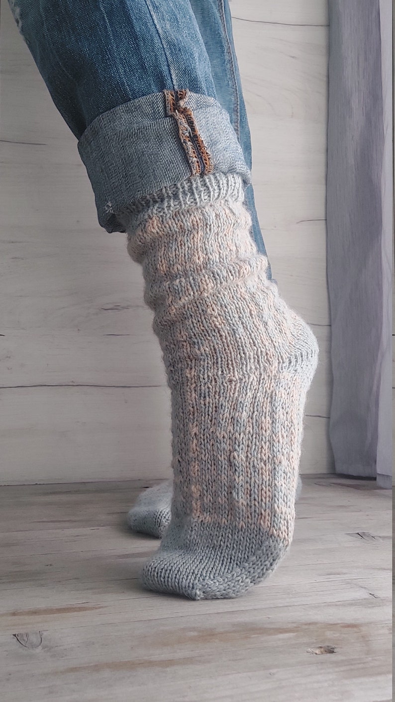 May include: A close-up of a pair of knitted socks in shades of light blue and peach. The socks are worn with blue jeans, with the cuffs of the jeans rolled up. The socks have a textured pattern.