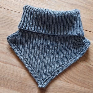 May include: A gray knitted cowl scarf with a ribbed collar and a triangular body. The scarf is made of a soft, textured yarn and is designed to be worn around the neck for warmth and style.