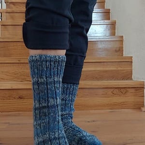 Puede incluir: Un par de calcetines de punto azul y blanco. Los calcetines se llevan con pantalones negros. Los calcetines tienen una textura acanalada y un patr&oacute;n moteado. El fondo muestra una escalera y un suelo de madera.