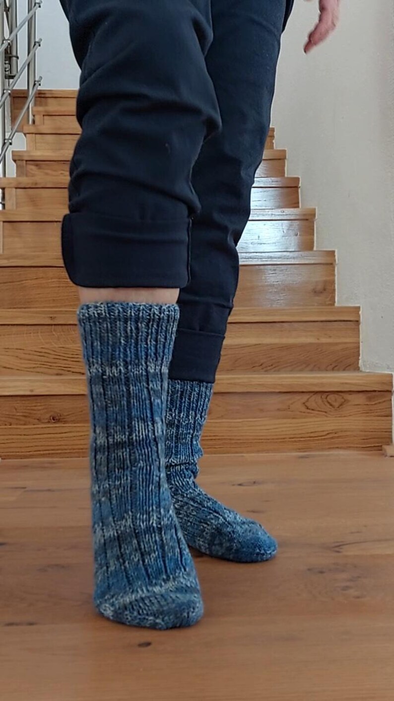 Puede incluir: Un par de calcetines de punto azul y blanco. Los calcetines se llevan con pantalones negros. Los calcetines tienen una textura acanalada y un patr&oacute;n moteado. El fondo muestra una escalera y un suelo de madera.