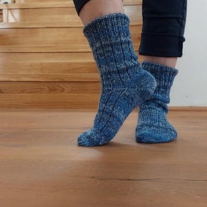 Puede incluir: Un par de calcetines azules de punto con un patr&oacute;n texturizado. Los calcetines se usan con pantalones azul oscuro. Los calcetines est&aacute;n sobre un suelo de madera, con una escalera de madera al fondo.
