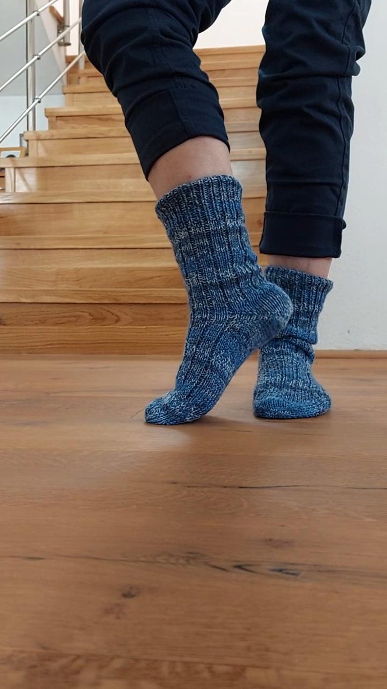 Puede incluir: Un par de calcetines azules de punto con un patr&oacute;n texturizado. Los calcetines se usan con pantalones azul oscuro. Los calcetines est&aacute;n sobre un suelo de madera, con una escalera de madera al fondo.